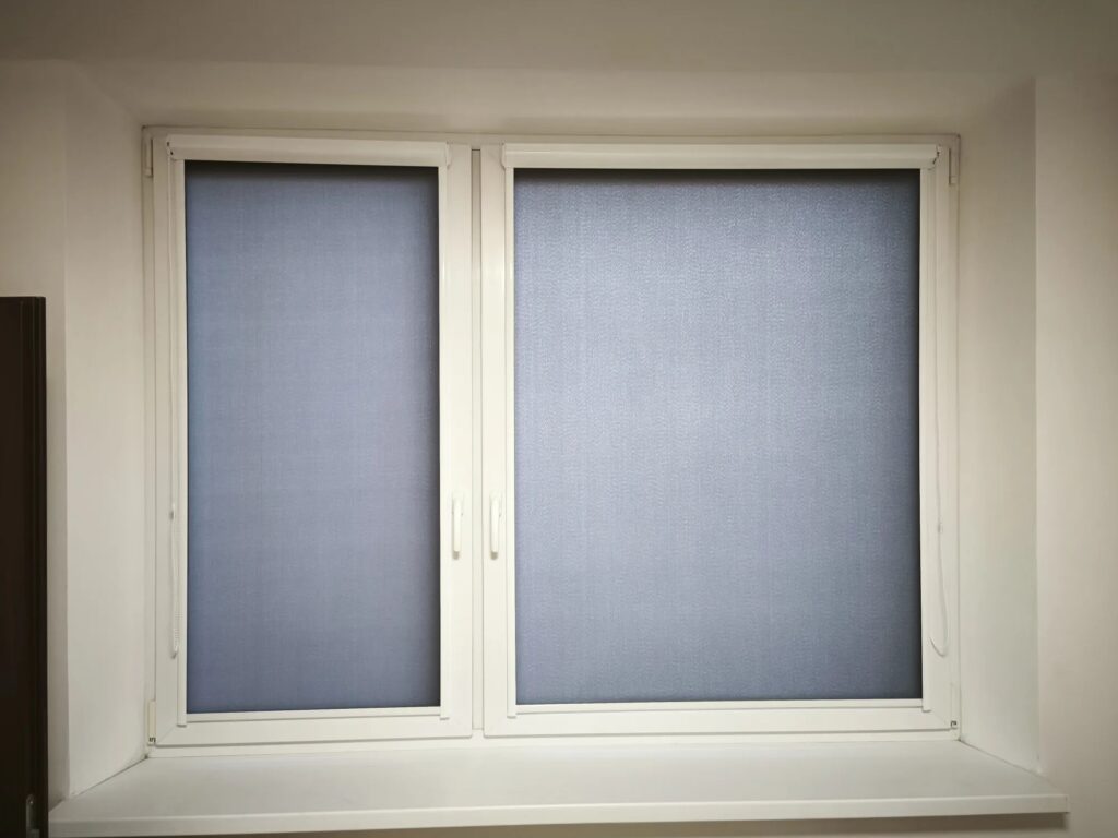Šedé zatemňovací rolety stažené na dvoukřídlém plastovém okně